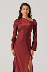 A woman is wearing a long, satin, burgundy-colored dress with long sleeves. The dress features a subtle draped detail at the neckline and a thigh-high slit on one side.